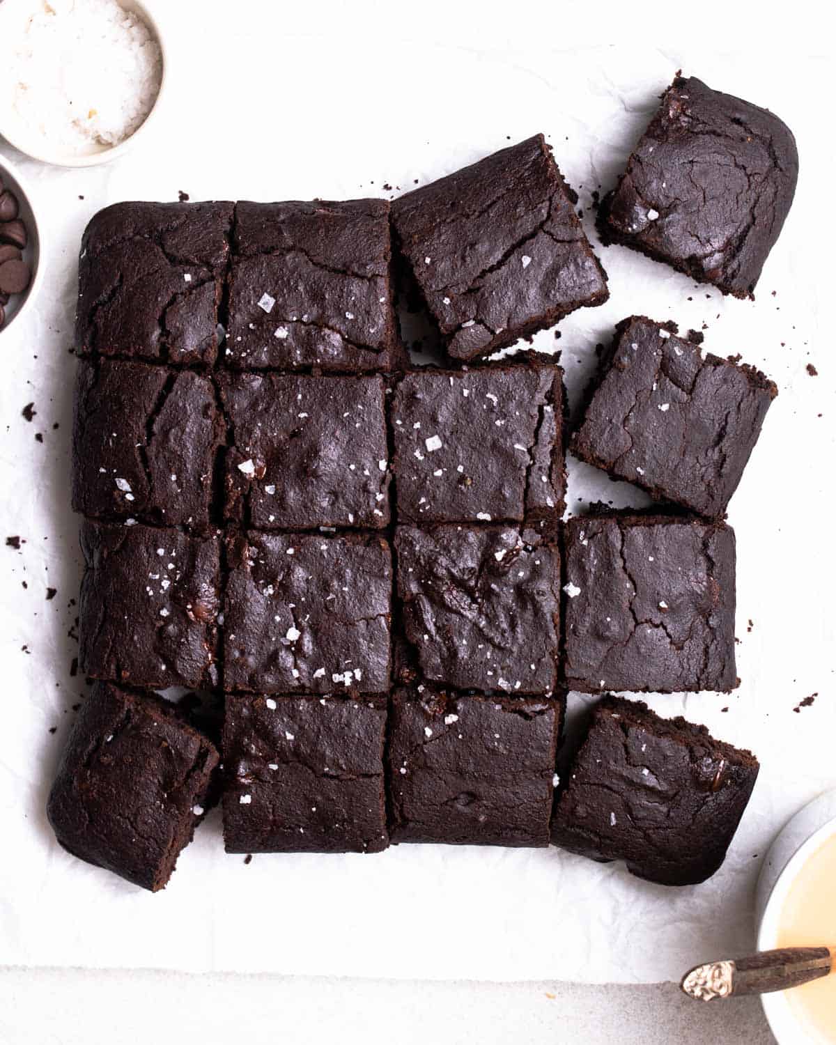 A tray of of sliced brownies arranged on a parchment paper with chocolate chips and flaked sea salt beside.