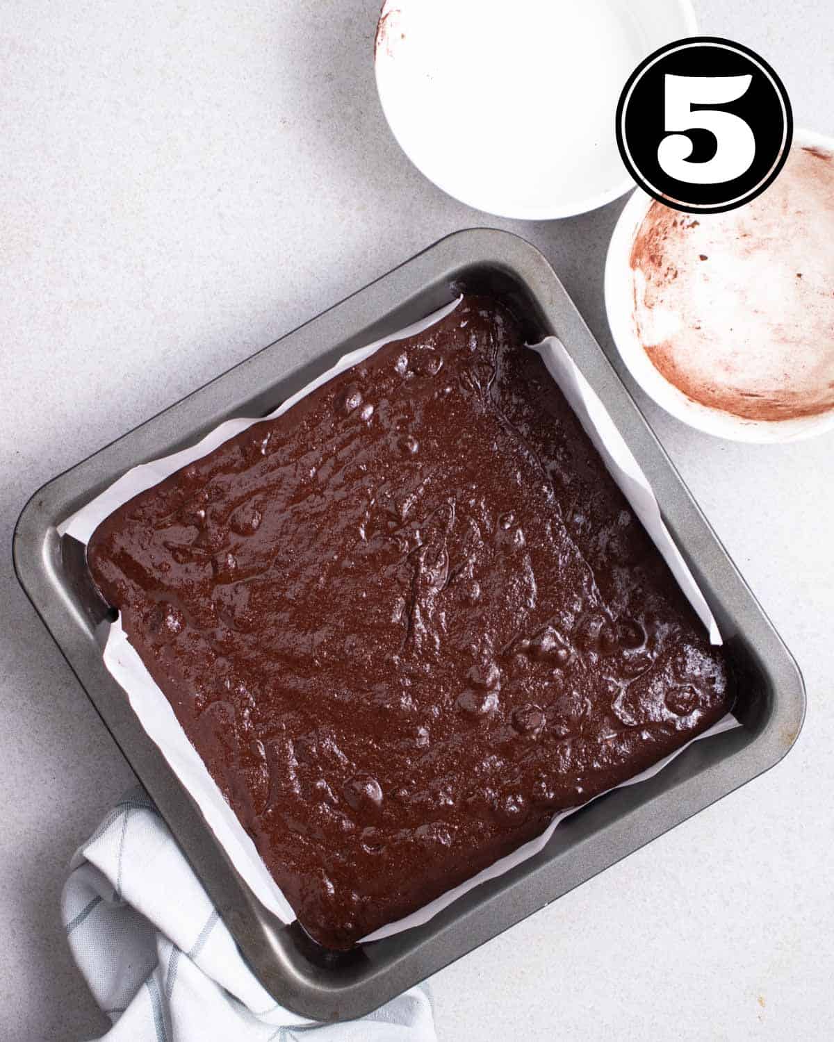 Brownie batter in a square baking tray lined with parchment paper with 2 bowls in the background.