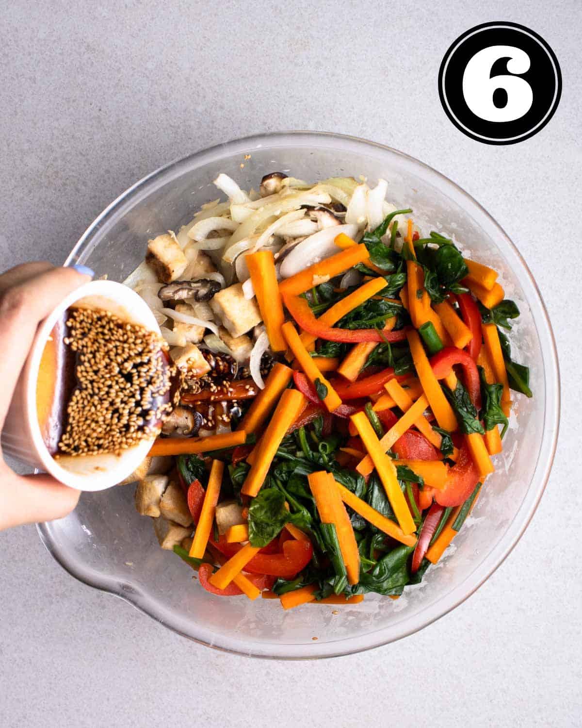Pouring japchae sauce into a bowl of cooked vegetables, noodles, and tofu in a glass bowl.