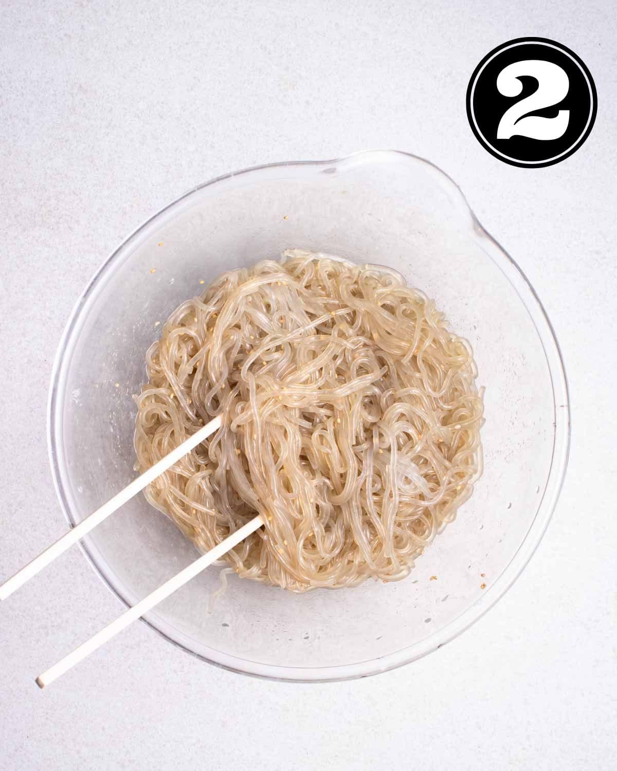 Cooked sweet potato noodles tossed in sauce in a glass bowl with a pair of chopsticks beside.