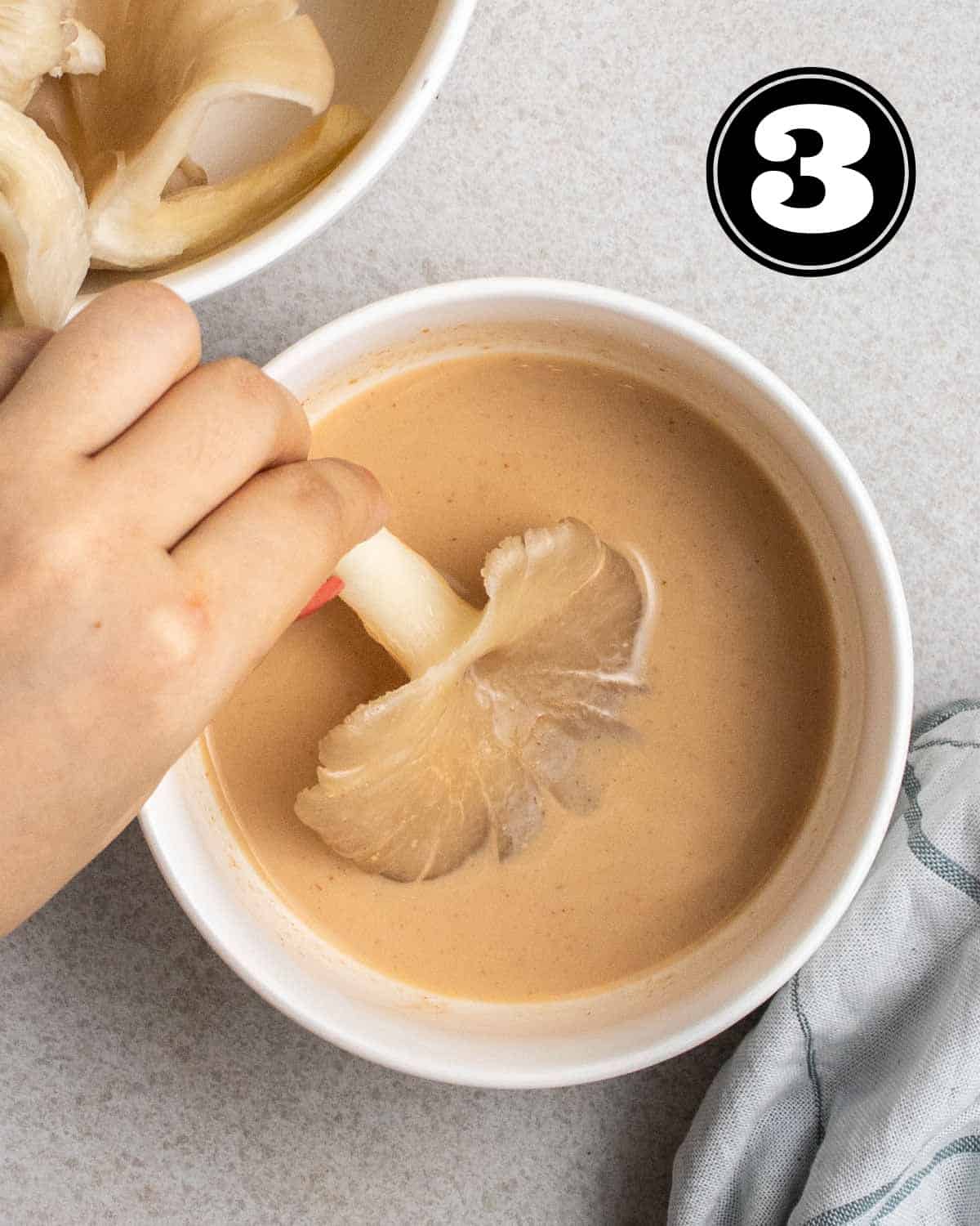 Dipping an oyster mushrooms in buttermilk with a bowl of oyster mushrooms beside.