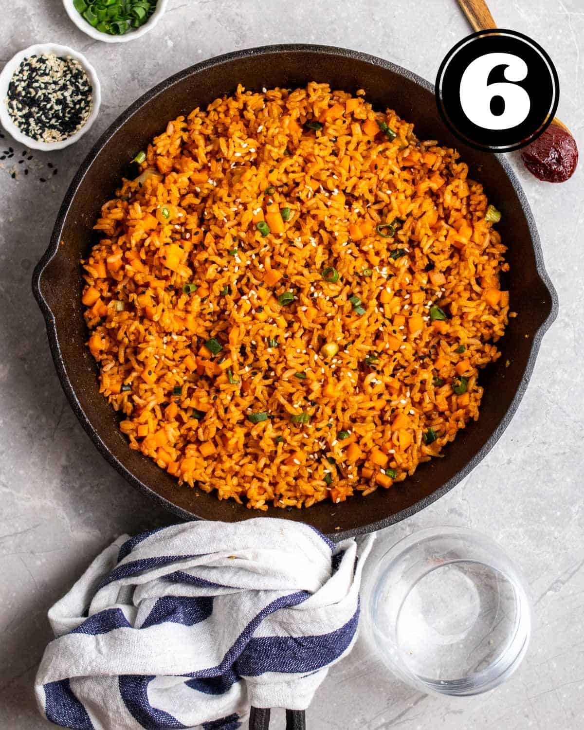 A pan of gochujang fried rice with spoonful of gochujang, sesame seeds, and chopped green onion beside.