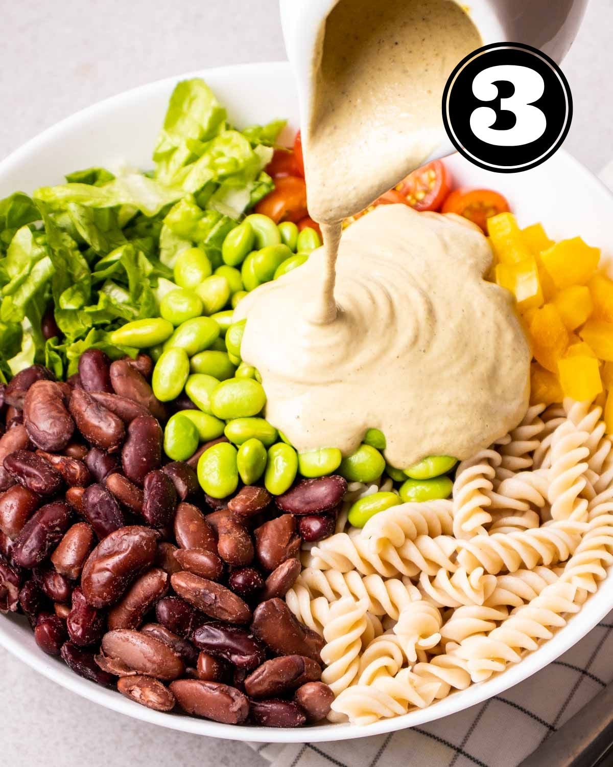Pouring dressing into a bowl of vegetables, beans, and cooked pasta.