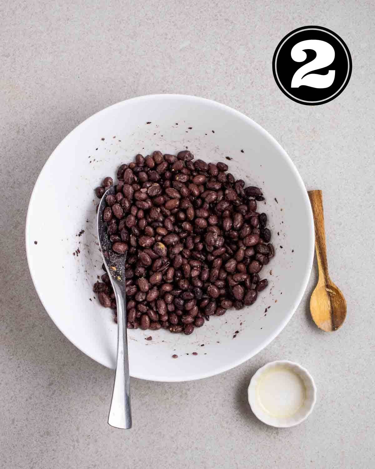 Seasoned black beans in a bowl with a spoon beside. There is a wooden spoon and sauce dish beside the bowl.