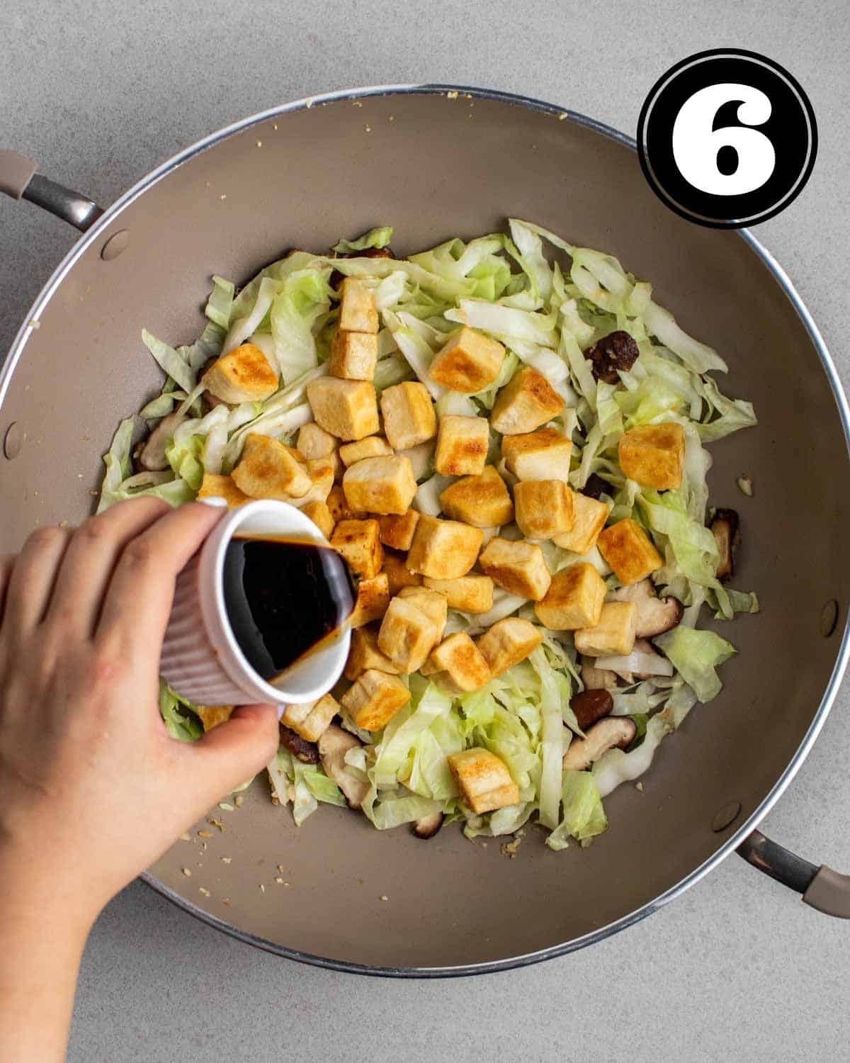 Pouring sauce into a wok of cooked cabbage, mushrooms, and pan-fried tofu.