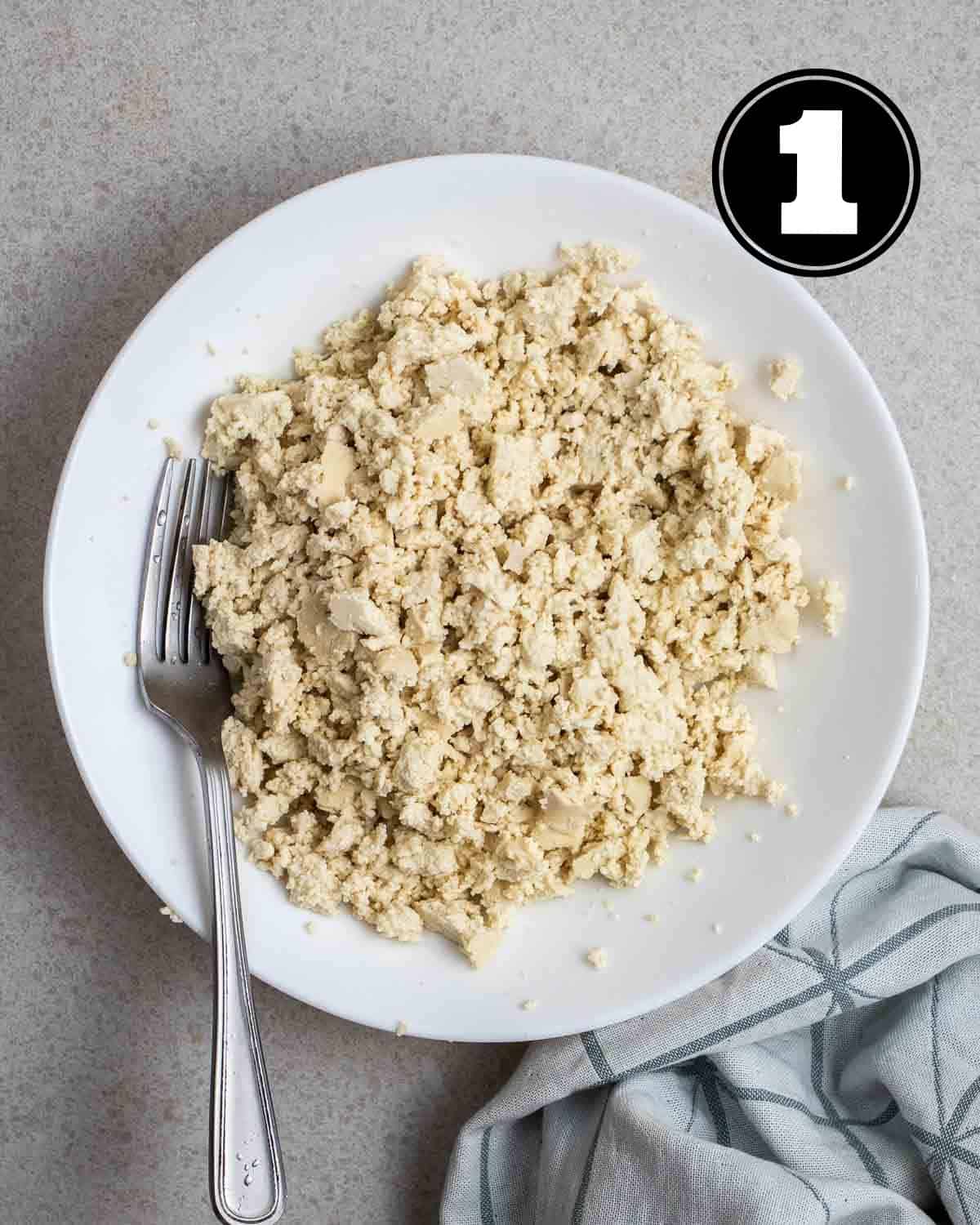 Crumbled tofu in a white plate with a fork beside with a blue teacloth beside.
