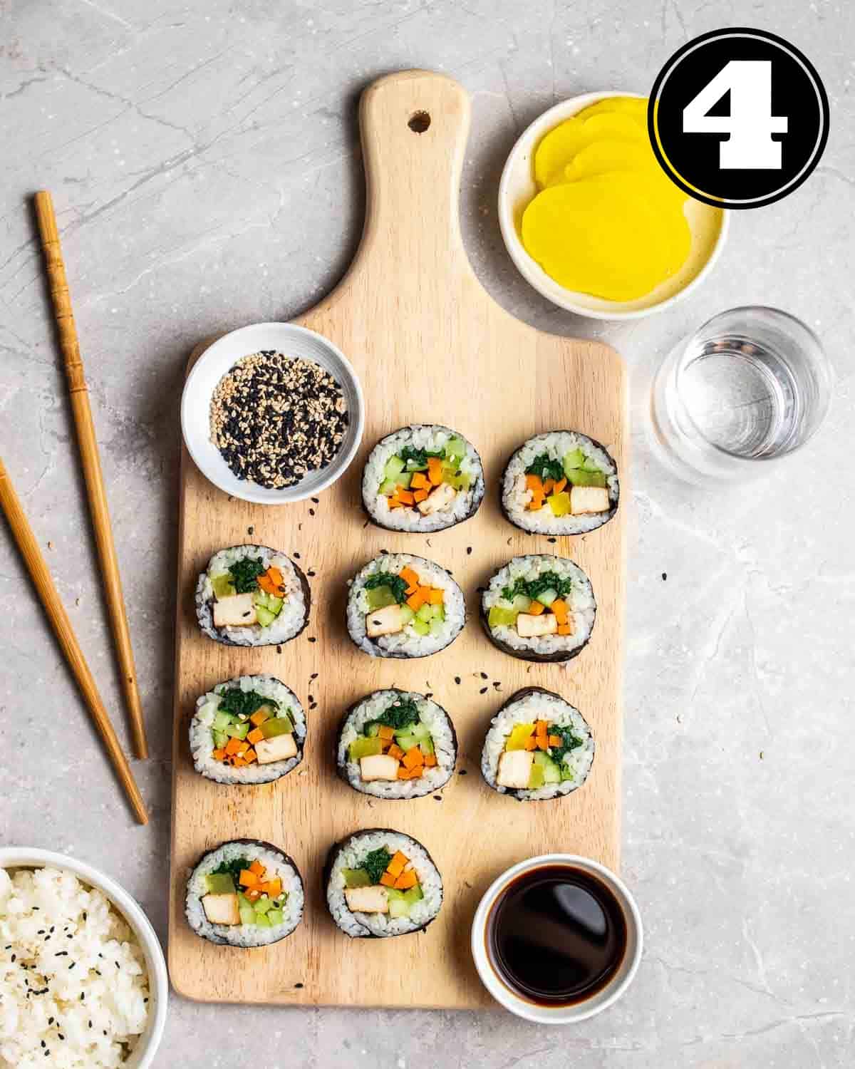 Slices of kimbap on a wooden board with a pair of chopsticks, damuji, soy sauce, water, and bowl of rice beside.