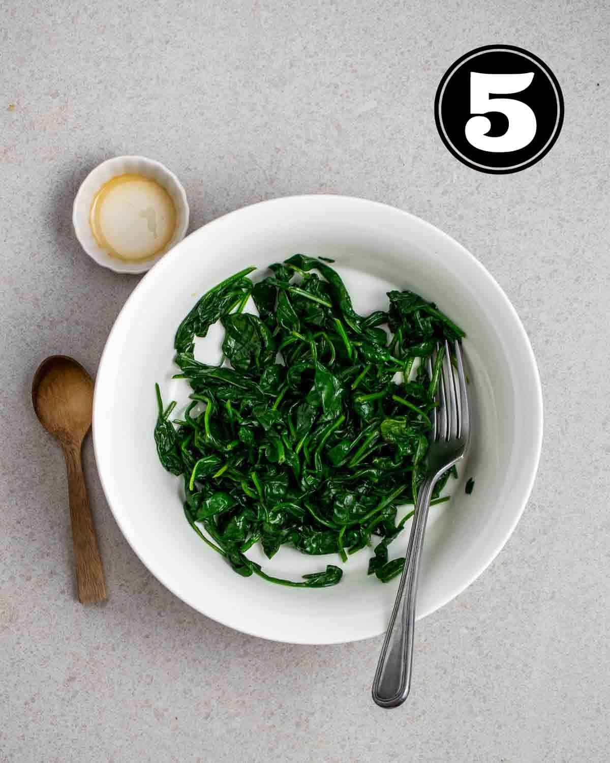 Seasoned, cooked spinach in a bowl with a fork beside and a wooden spoon and sauce dish in the background.