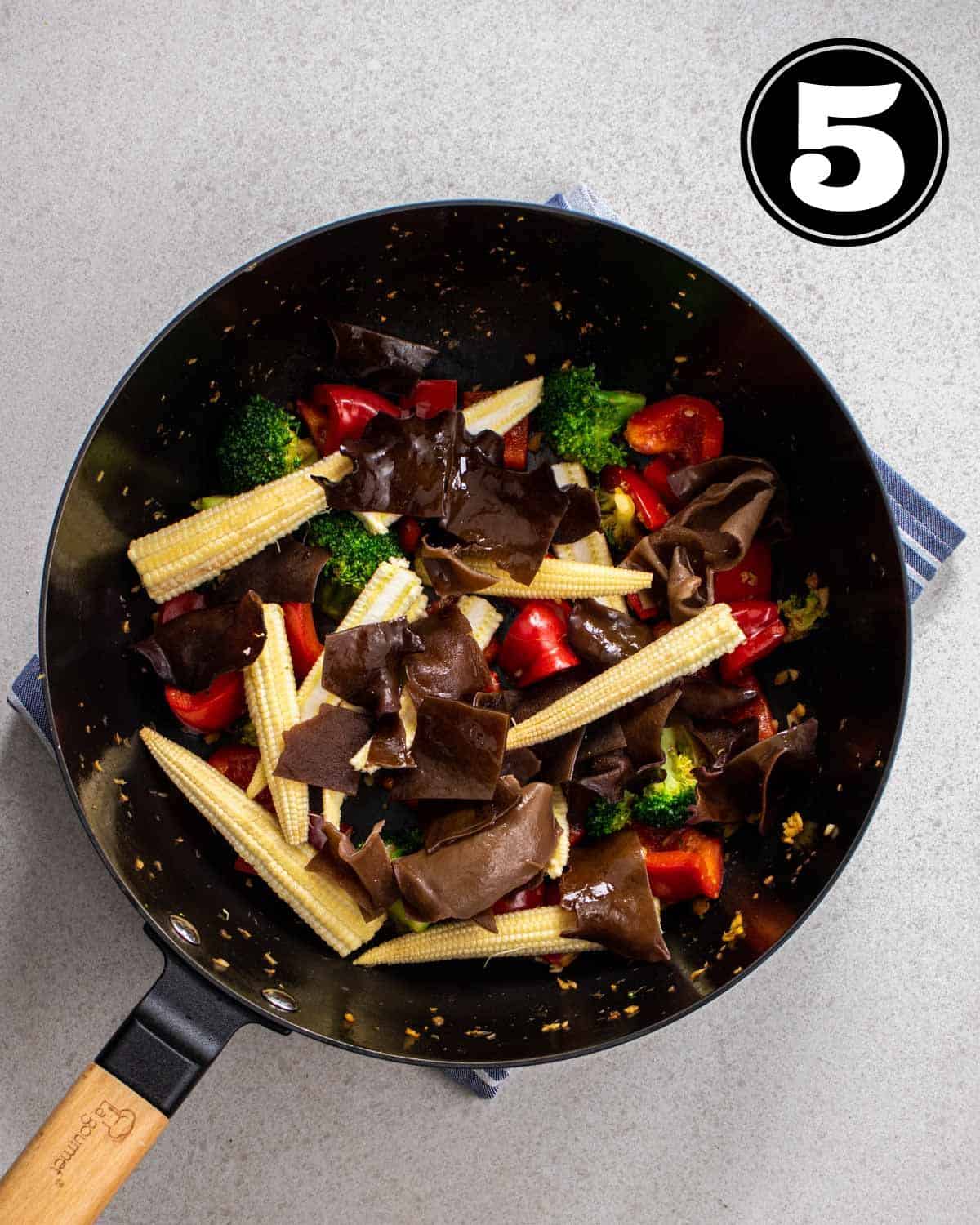A wok of cooked vegetables in a cast iron wok on a blue folded teacloth.