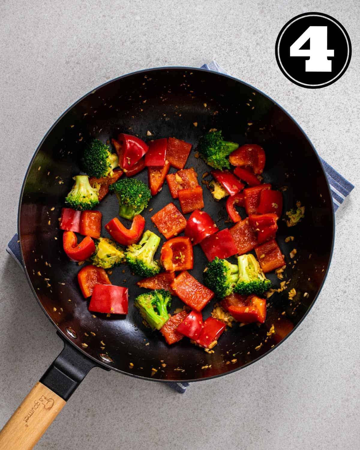 Cooked broccoli and bell pepper in a cast iron wok on a blue and white striped teacloth.