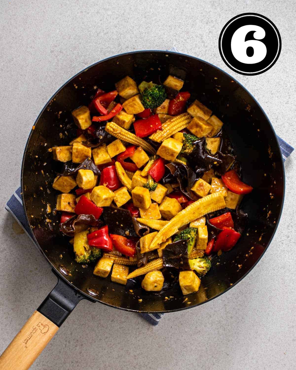 A wok of tofu and vegetables on a blue and white striped teacloth.