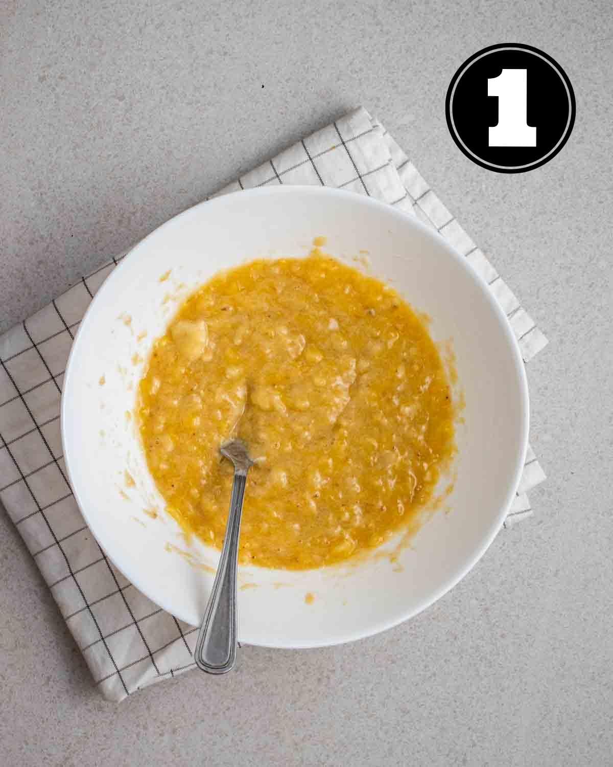 Mashed bananas in a white bowl with a fork beside on a white folded checkered teacloth.