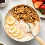 A bowl of yogurt topped with granola and banana slices with a spoon beside.