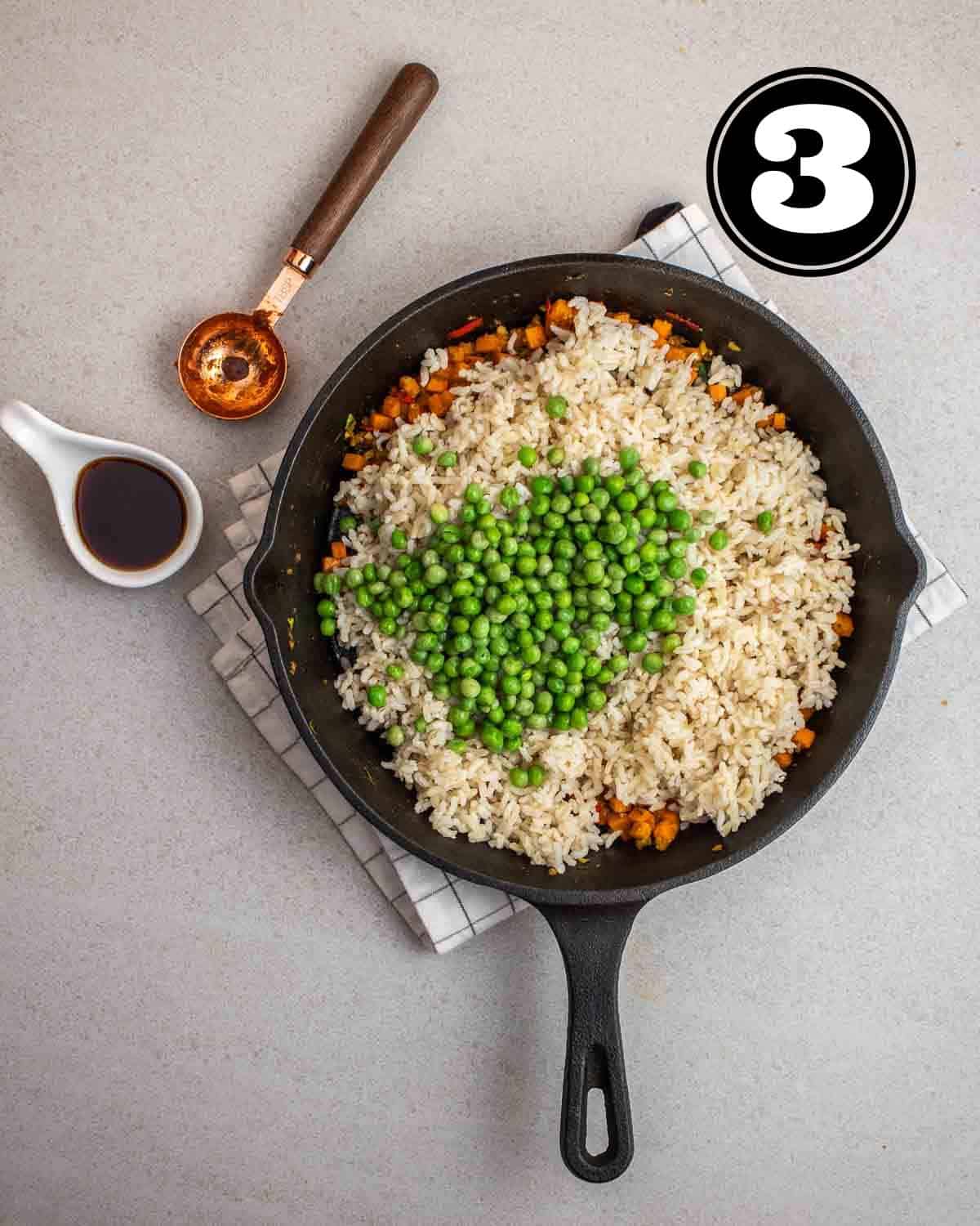 Peas and rice added to cooked carrot in a skillet on a white folded teacloth.