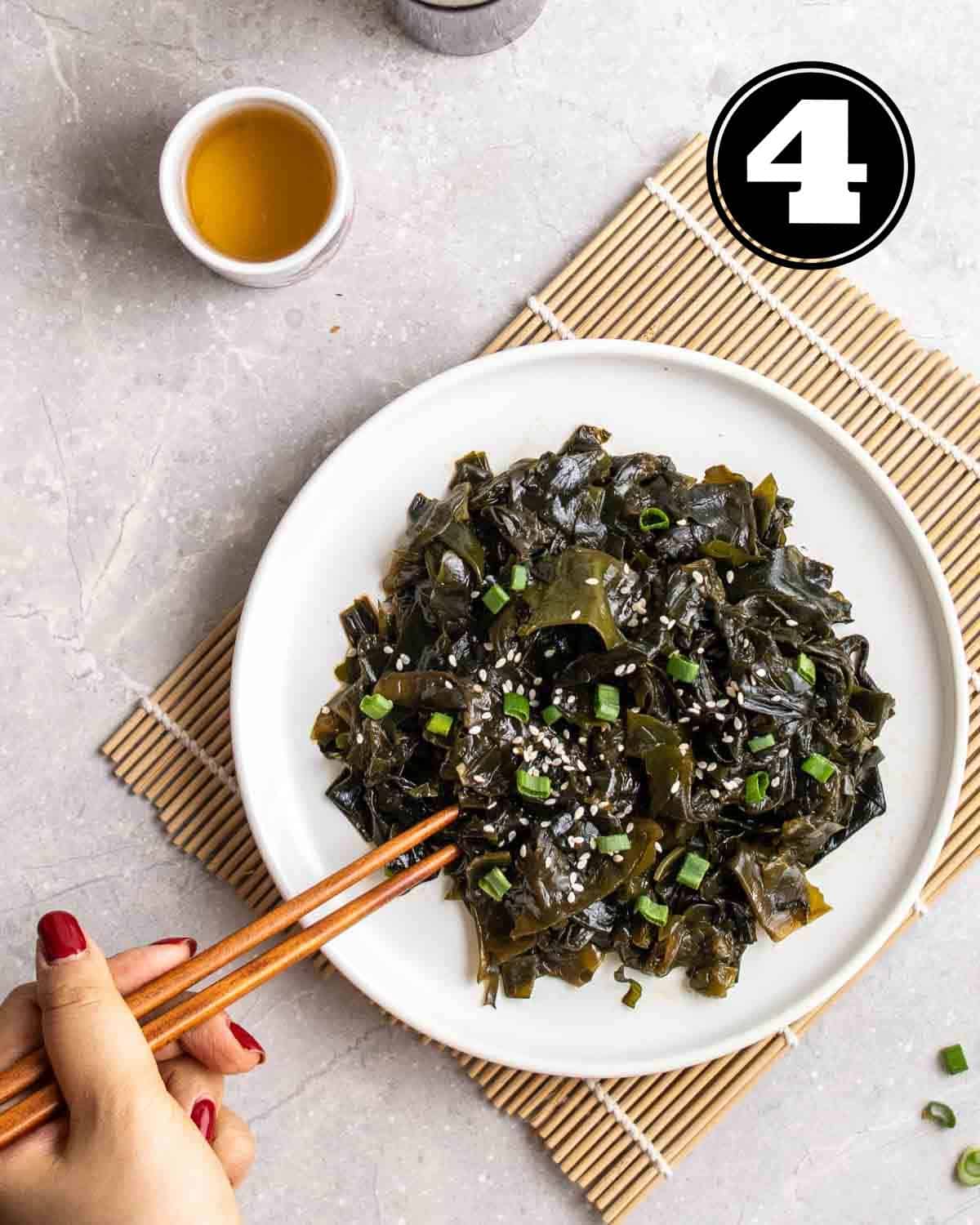 Stirring a plate of wakame salad with a pair of chopsticks on a bamboo sushi mat with a cup of tea beside.