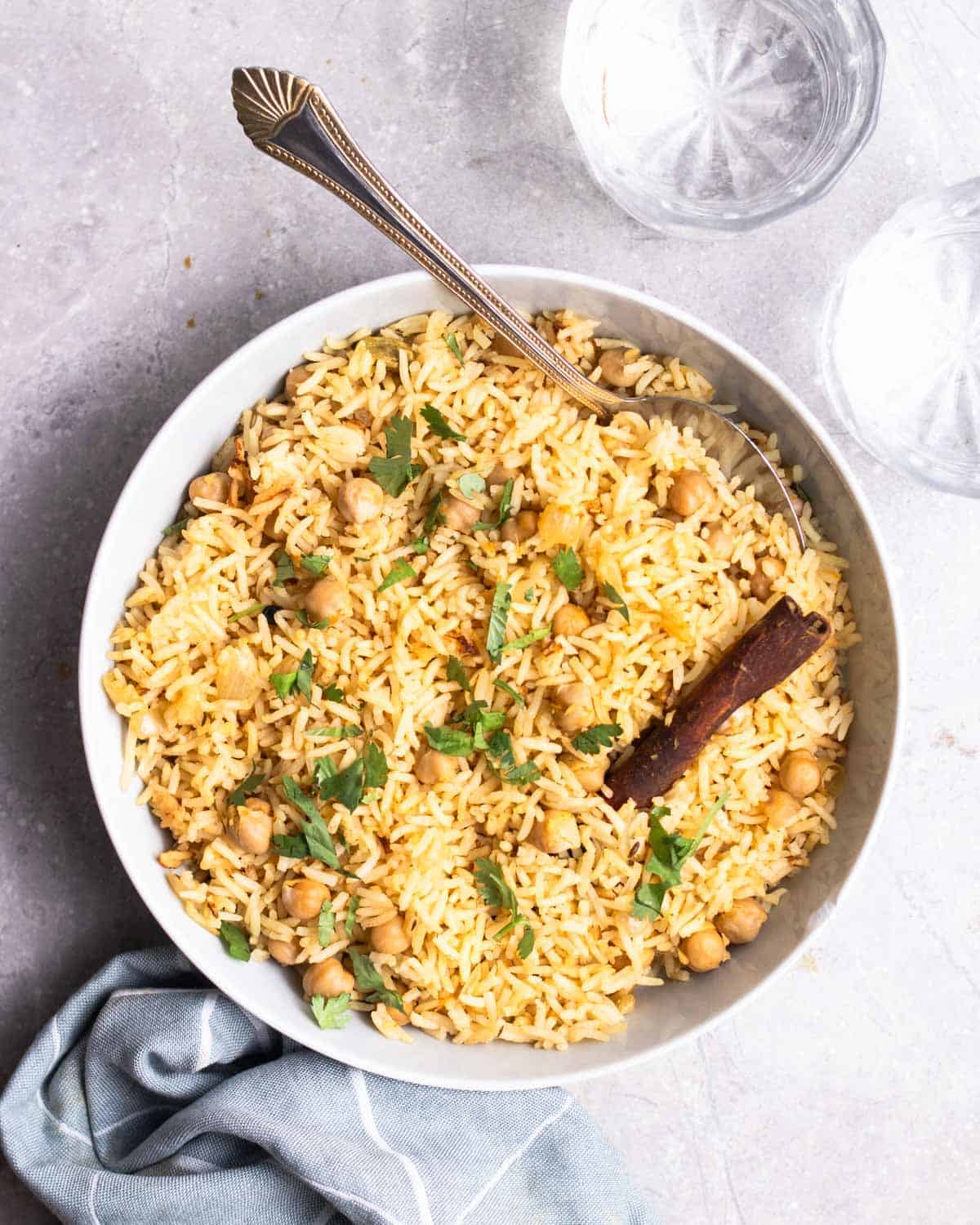 A plate of chickpea pulao served with spoon with 2 glasses of water and a blue teacloth in the background.