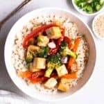 Teriyaki tofu served on a bowl of rice with a fork, chopped green onion, and sesame seeds beside.
