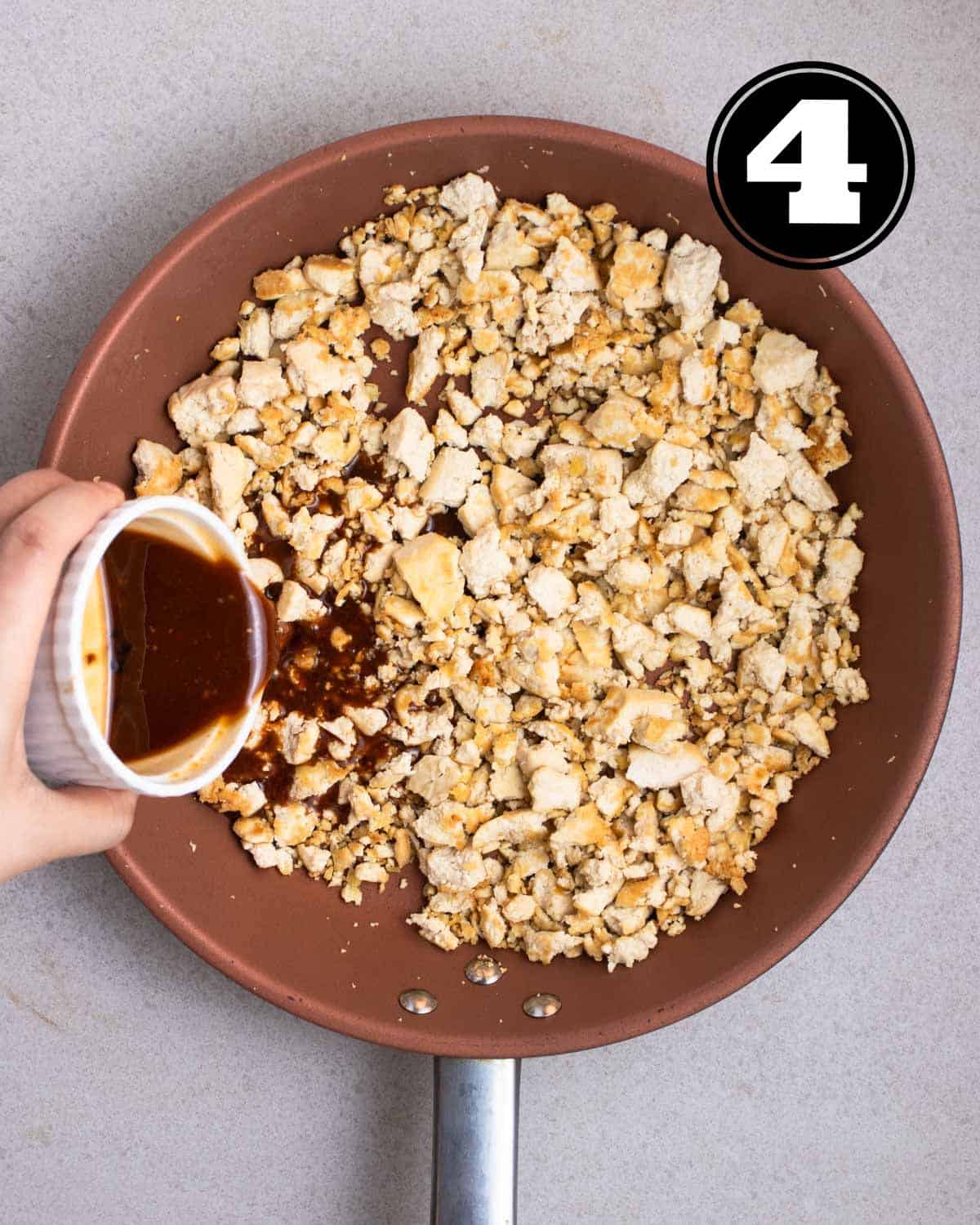 Pouring stir-fry sauce into a pan of cooked crumbled tofu in a non-stick pan.