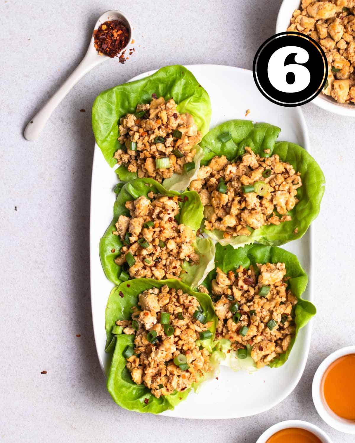 A plate of tofu lettuce wraps with a spoonful of chili flakes, 2 cups of tea and bowl of tofu mixture beside.