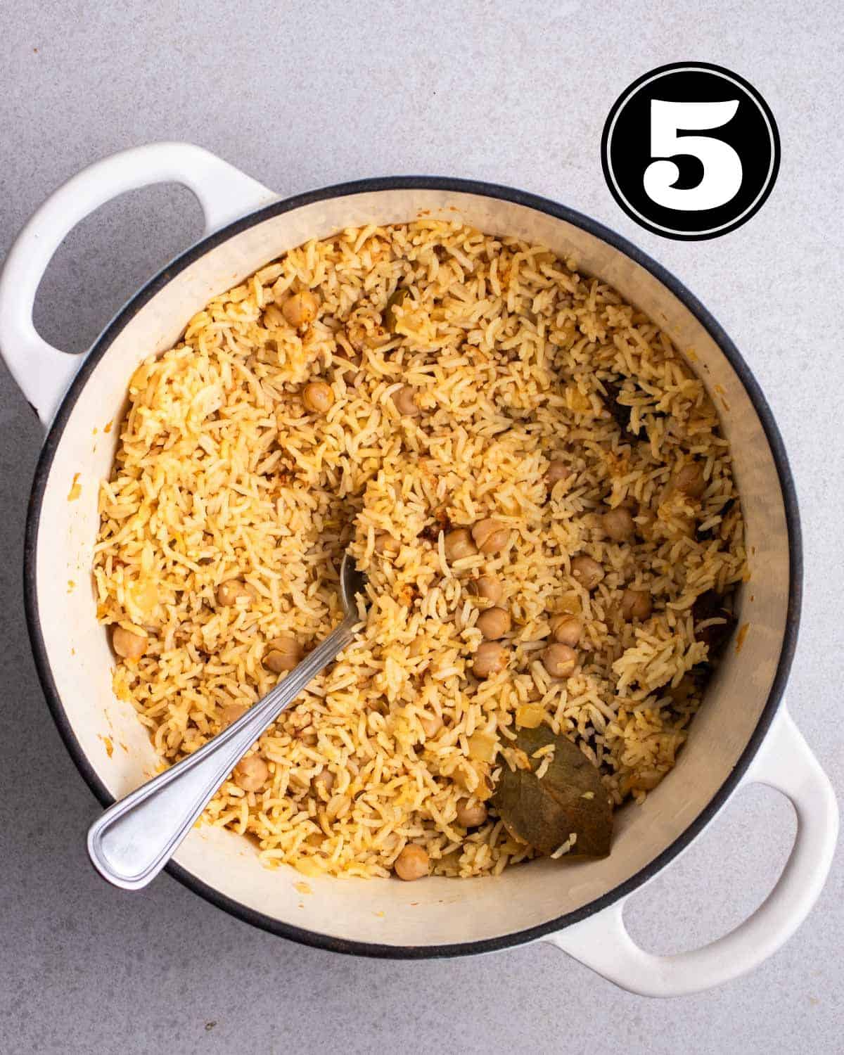 Cooked chickpea and rice in a white Dutch oven with a spoon beside.