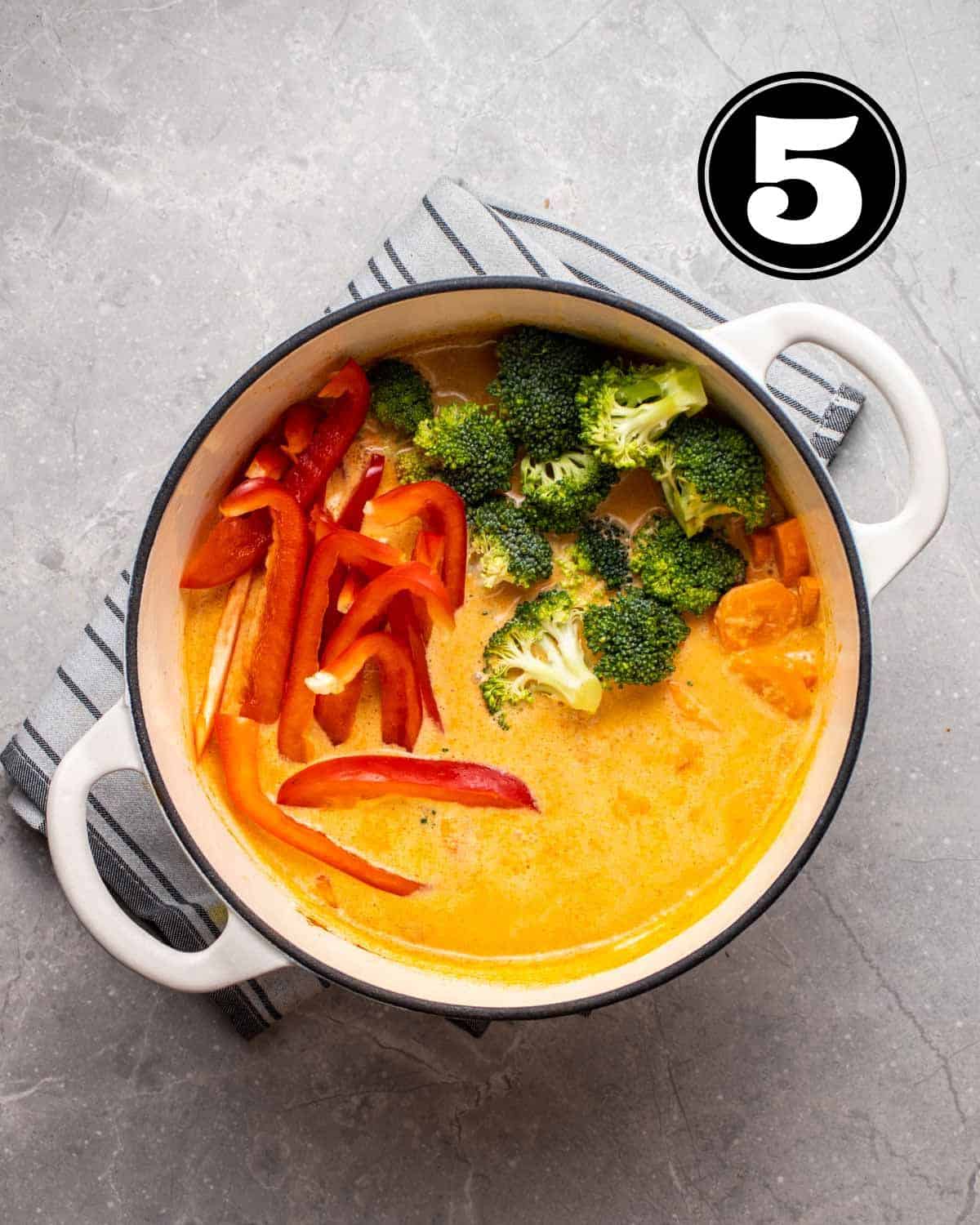 Uncooked vegetables in a red curry sauce in a white Dutch oven.