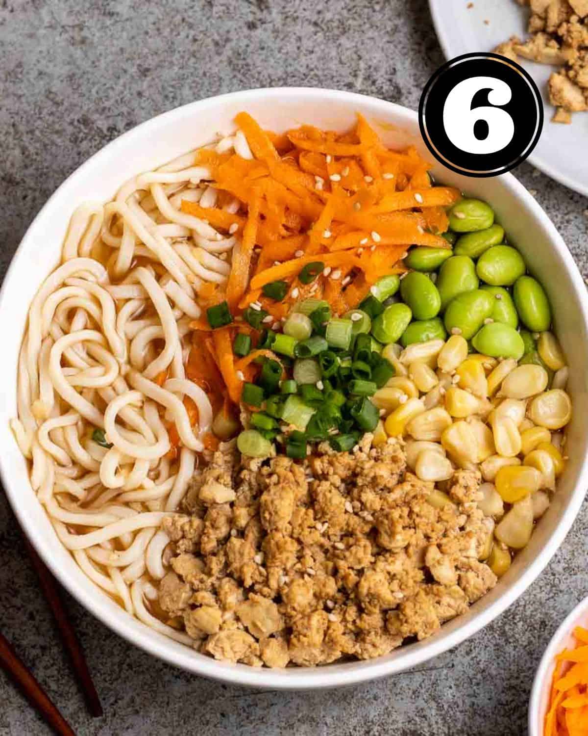 A bowl of ramen topped with edamame, corn, shredded carrot, green onion, and minced tofu.