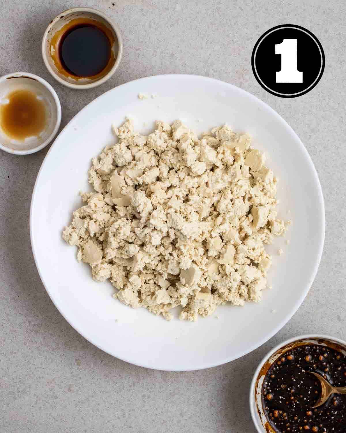 Crumbled up tofu in a white plate with 2 sauce dishes and ramekin with sauce beside.