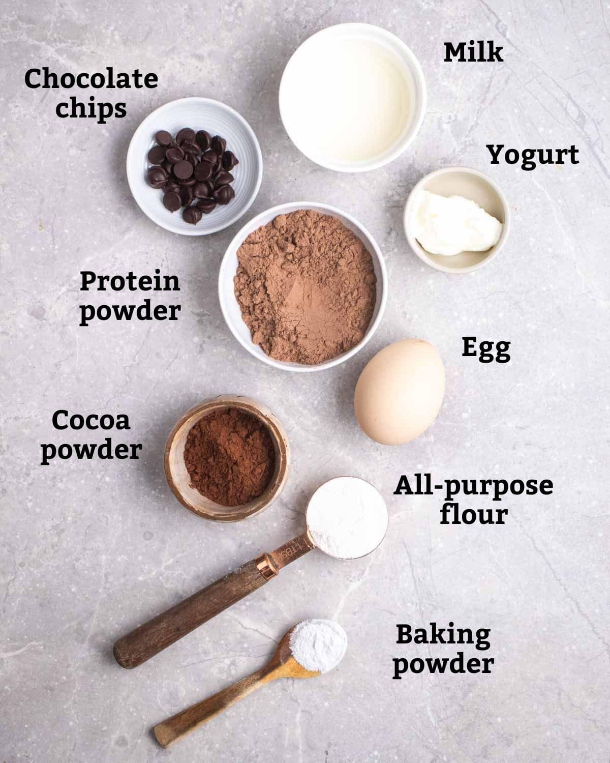 Ingredients needed for protein mug cake such as chocolate chips, milk, protein powder, egg, yogurt, cacao powder, flour, and baking powder.