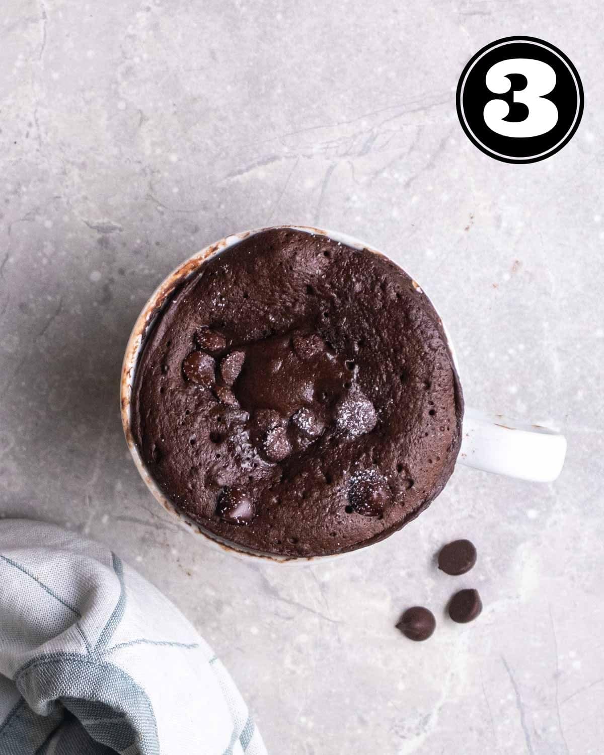 Cooked mug cake topped with chocolate chips with a blue teacloth and chocolate chips in the background.