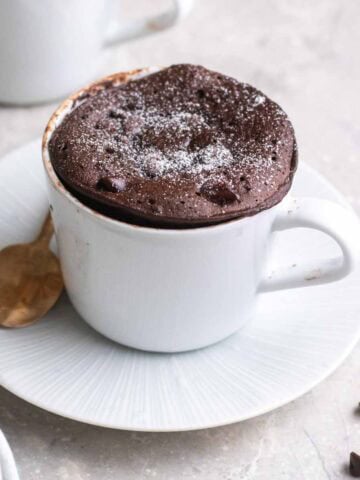 Protein mug cake topped with icing sugar served on saucer with a teaspoon beside.
