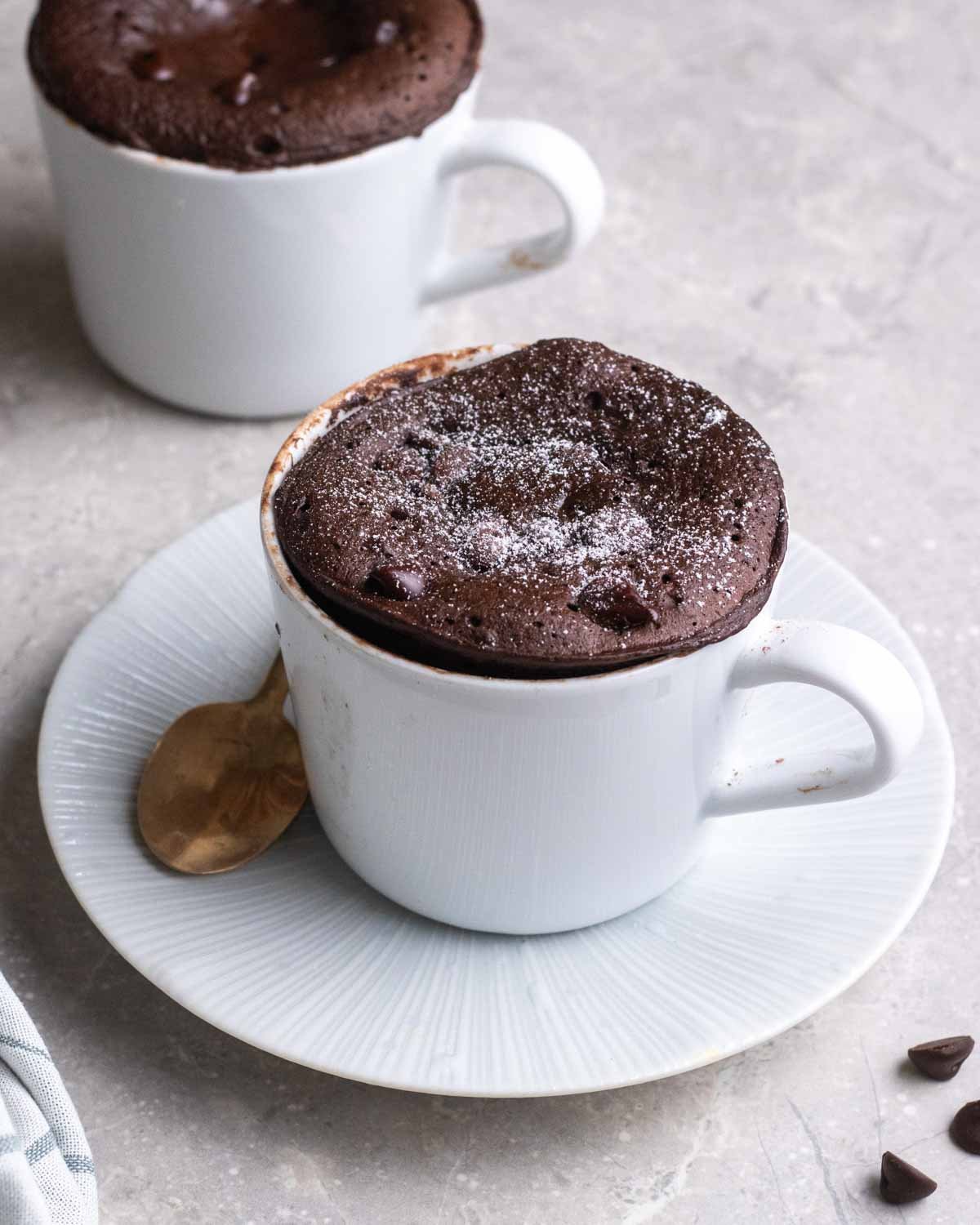 2 mugs of protein mug cake, which one of them on a saucer served with spoon.