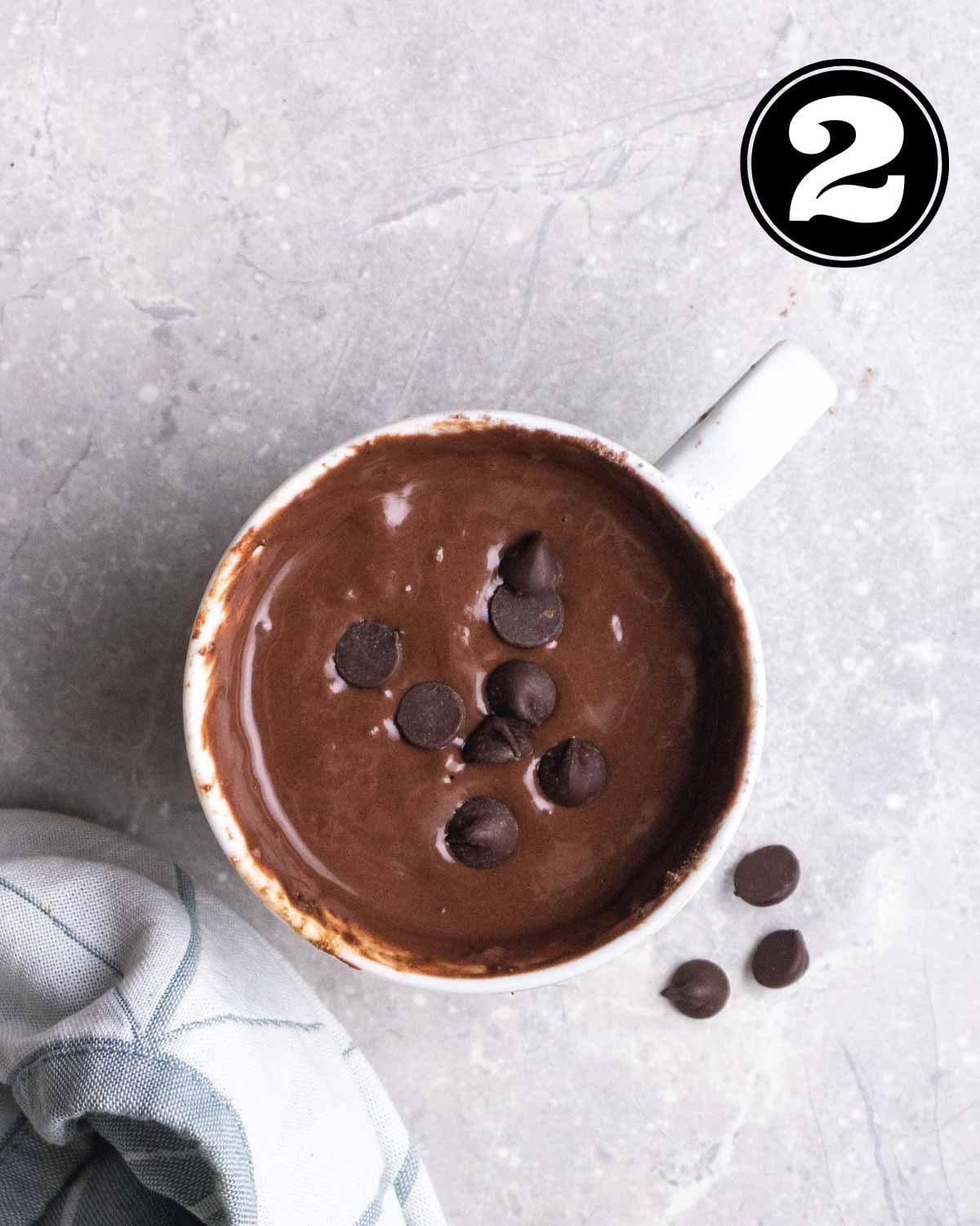 Batter of protein mug cake in a mug with chocolate chips and a blue teacloth beside.